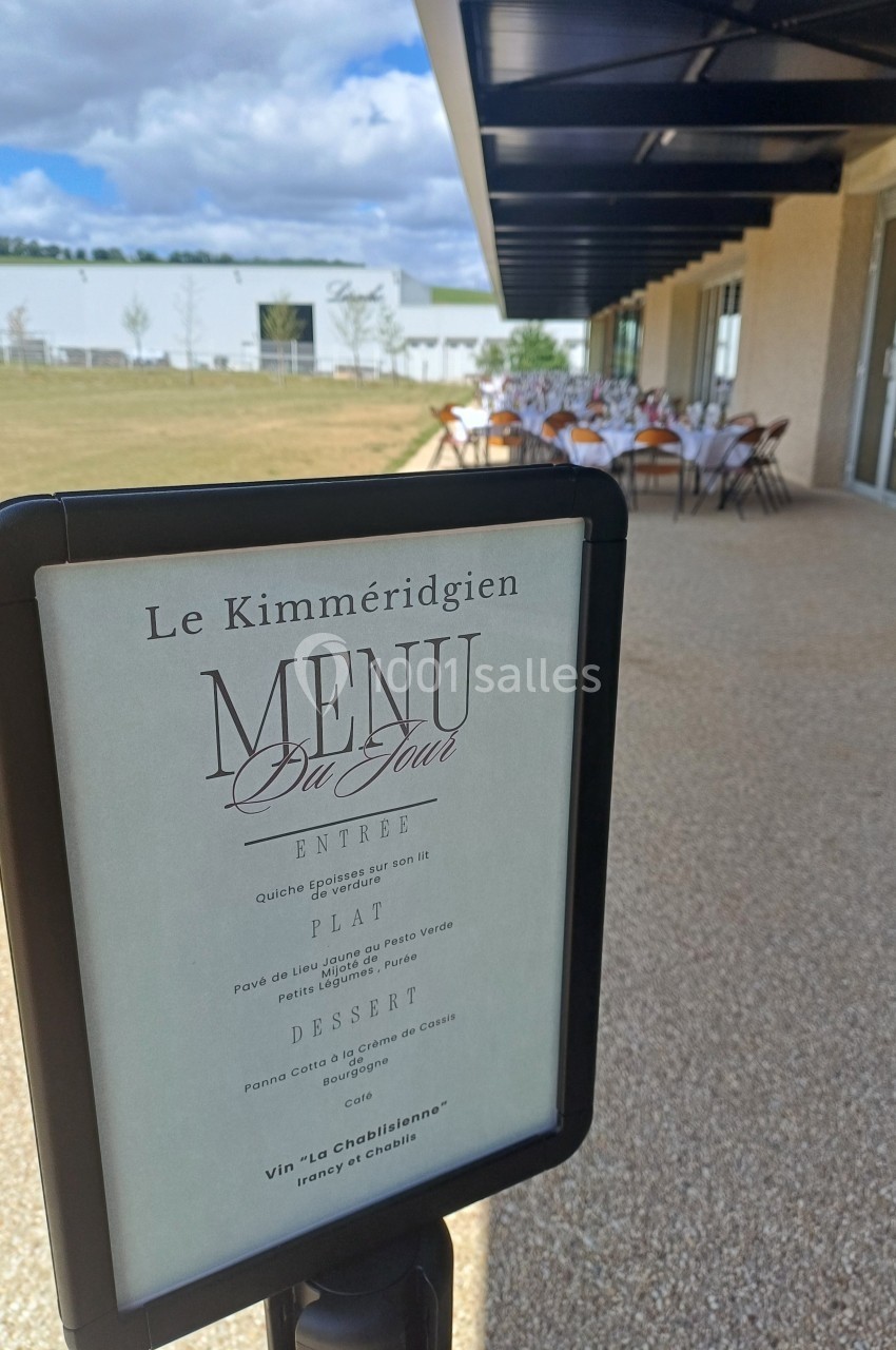 Panneau affichant le menu du jour d'un restaurant, avec une terrasse dressée en arrière-plan.