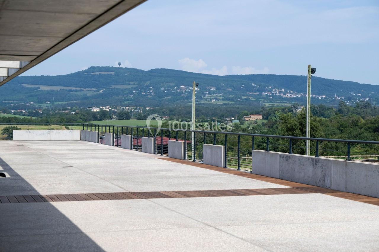 Vue d'une terrasse en béton avec garde-corps, donnant sur un paysage vallonné et verdoyant sous un ciel dégagé.