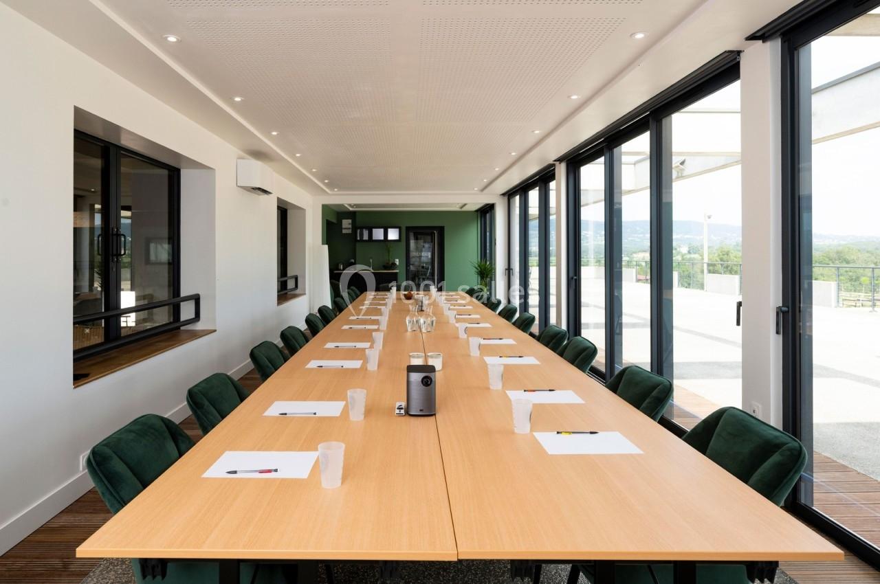 Salle de réunion lumineuse avec une grande table en bois, des chaises vertes et des baies vitrées donnant sur l'extérieur.