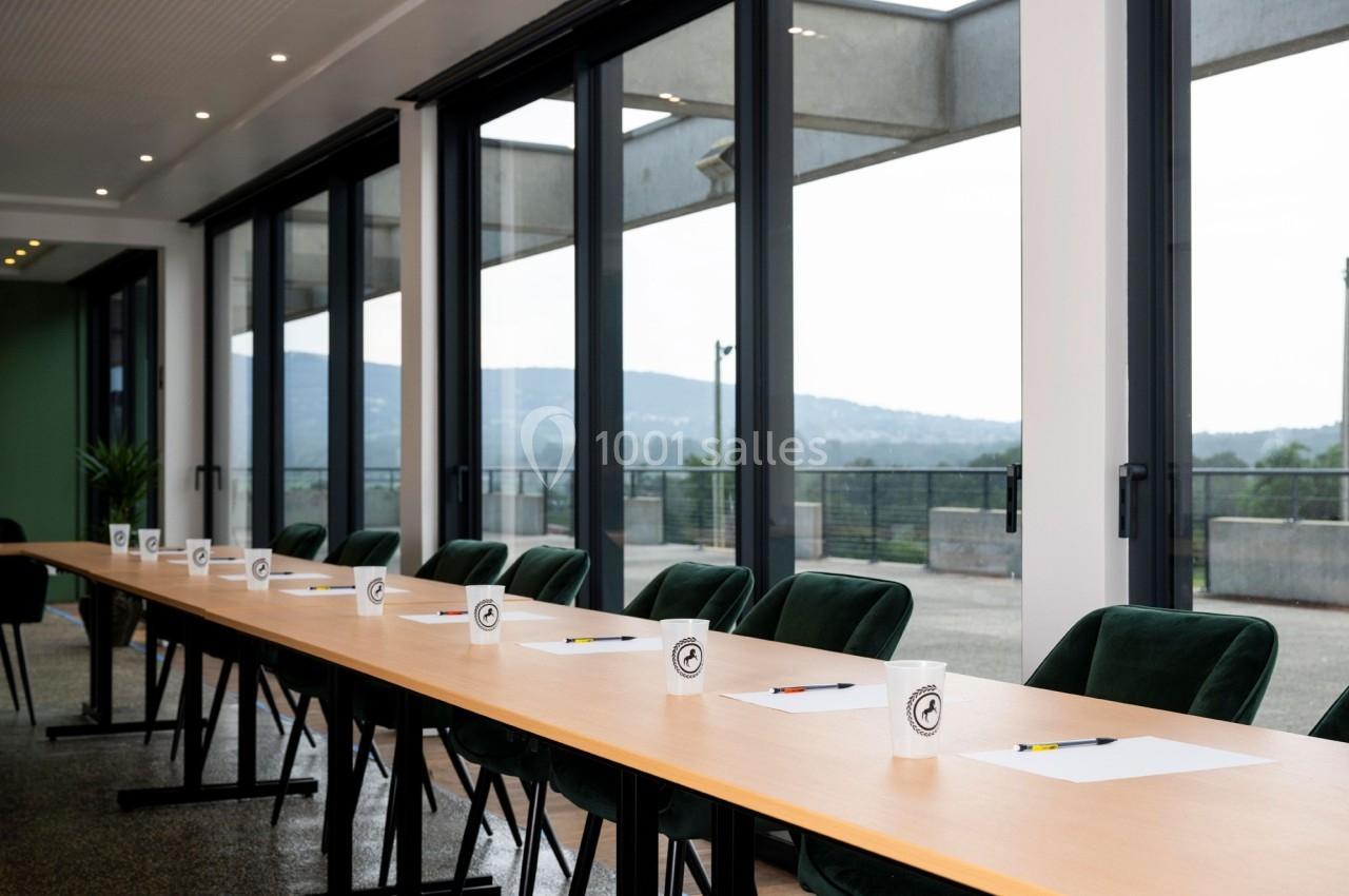 Salle de réunion lumineuse avec une longue table, des chaises vertes, des blocs-notes, des stylos et des gobelets alignés.