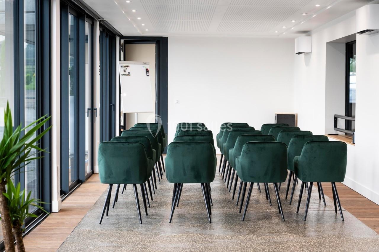 Salle lumineuse avec des chaises vertes alignées face à un tableau blanc, près de grandes baies vitrées.