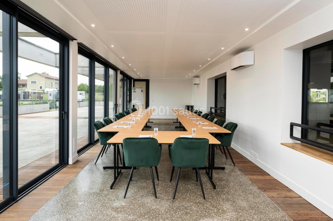 Salle de réunion lumineuse avec de grandes baies vitrées, tables en bois et chaises vertes alignées.