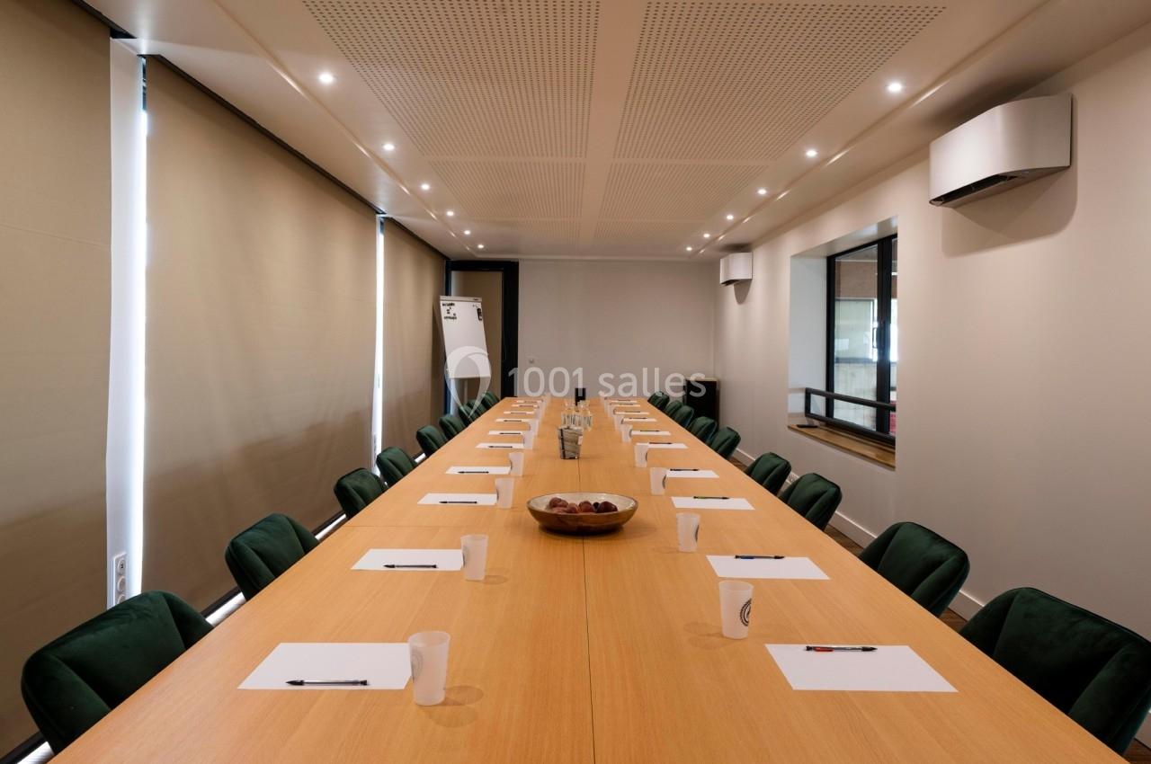 Salle de réunion avec une grande table en bois, chaises vertes, blocs-notes, stylos et carafe d'eau au centre.