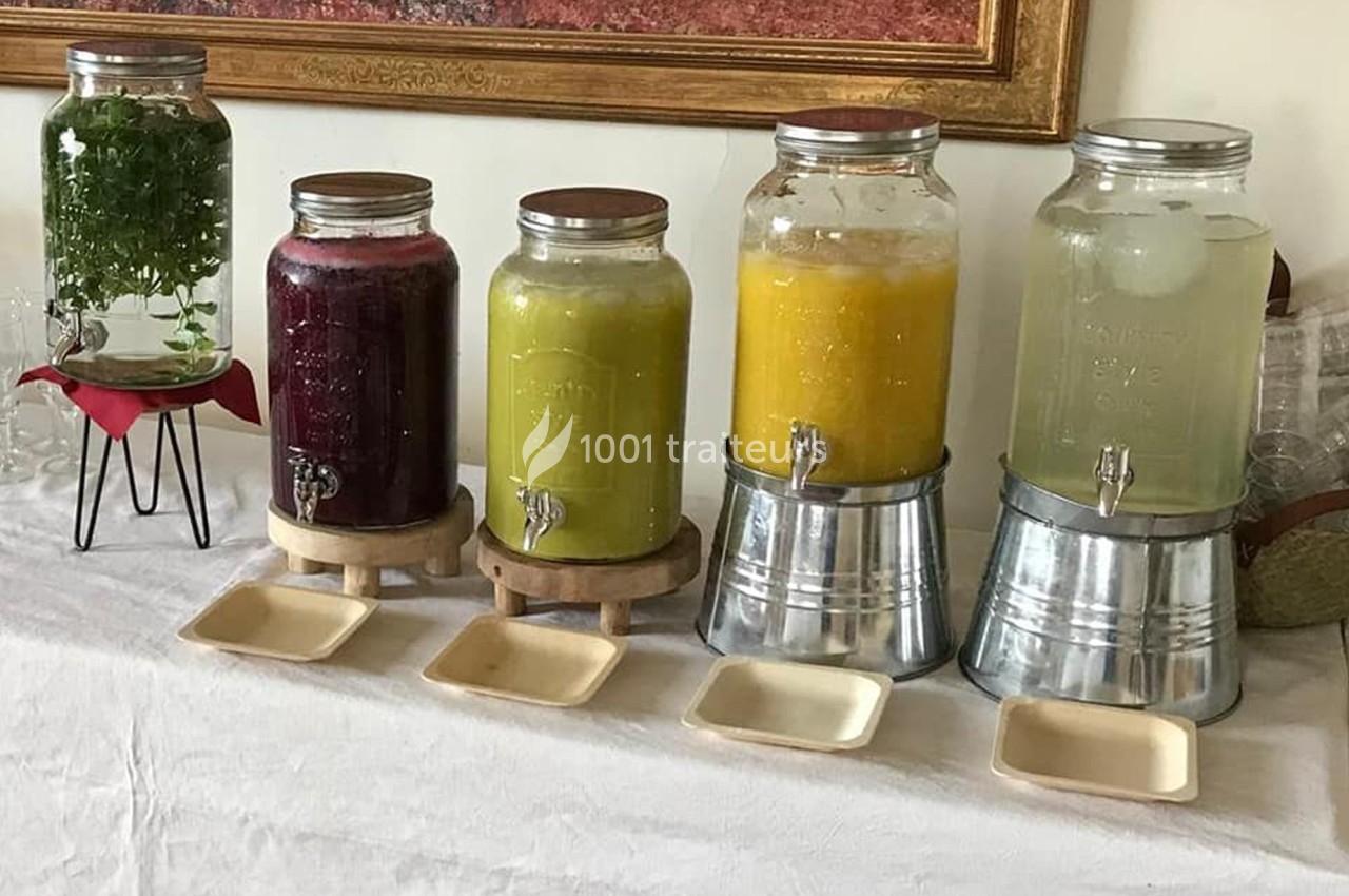 Quatre distributeurs en verre contenant des boissons colorées et des herbes, alignés sur une table blanche.