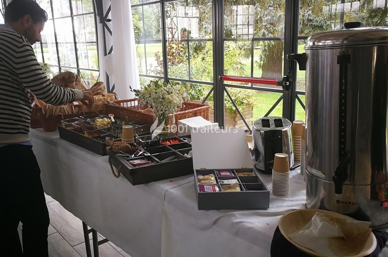 Un homme prépare un buffet avec boissons chaudes, viennoiseries et condiments, près d'une grande baie vitrée.