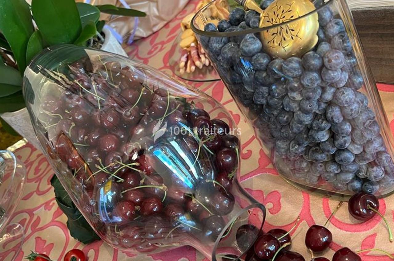 Deux vases en verre remplis de cerises rouges et de raisins noirs, posés sur une nappe rose à motifs.