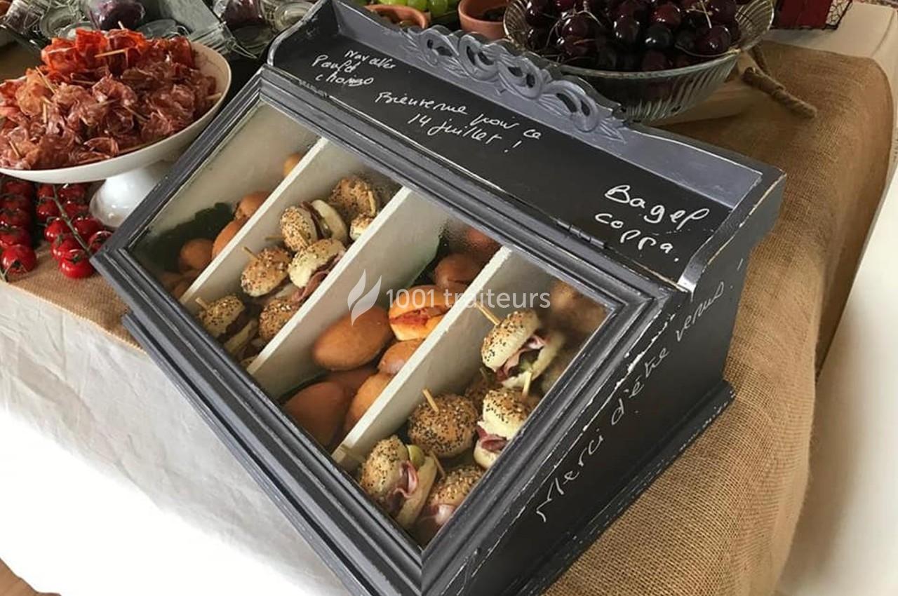 Présentoir en bois contenant des bagels et des pains, entouré de fruits, charcuteries et autres aliments sur une table.