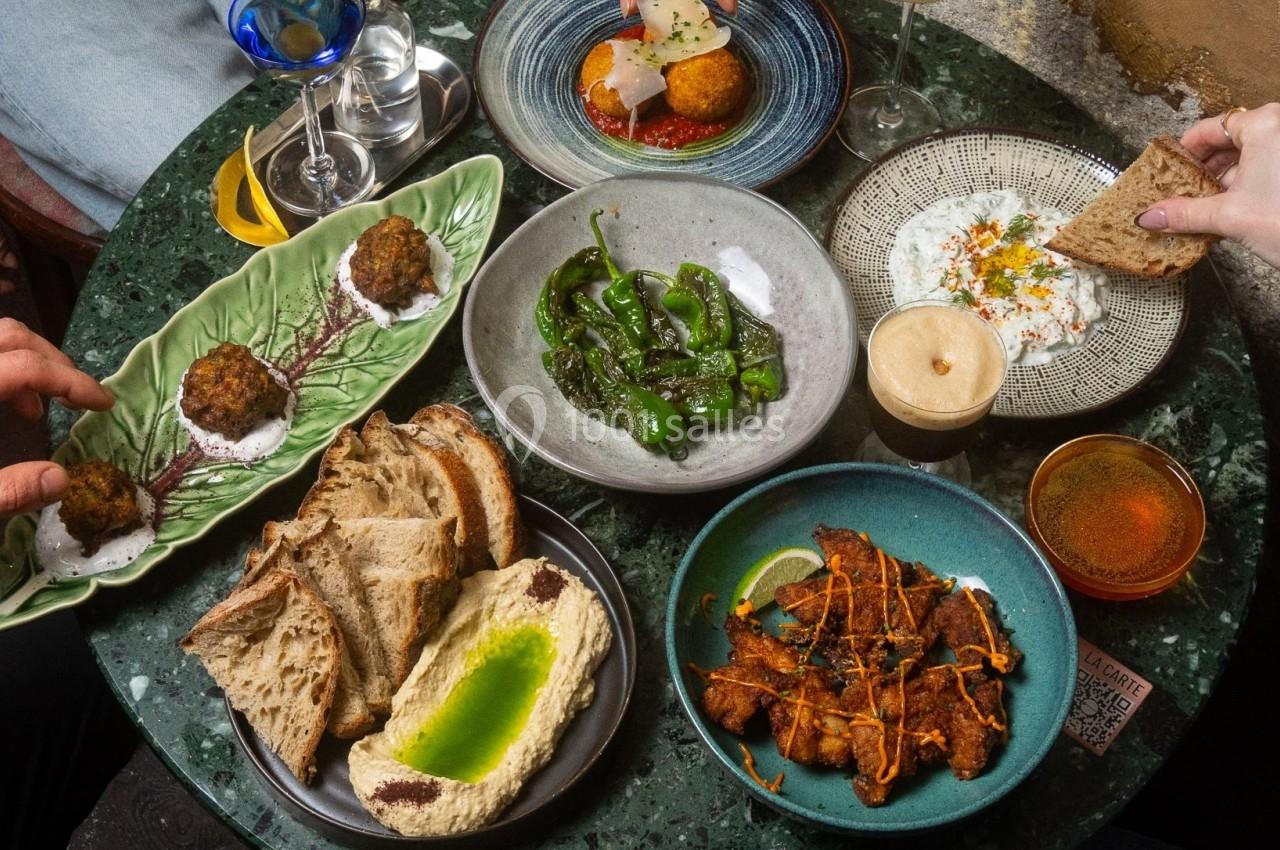 Table garnie de plats variés : pain, houmous, légumes, tapas et boissons, avec des mains en train de se servir.