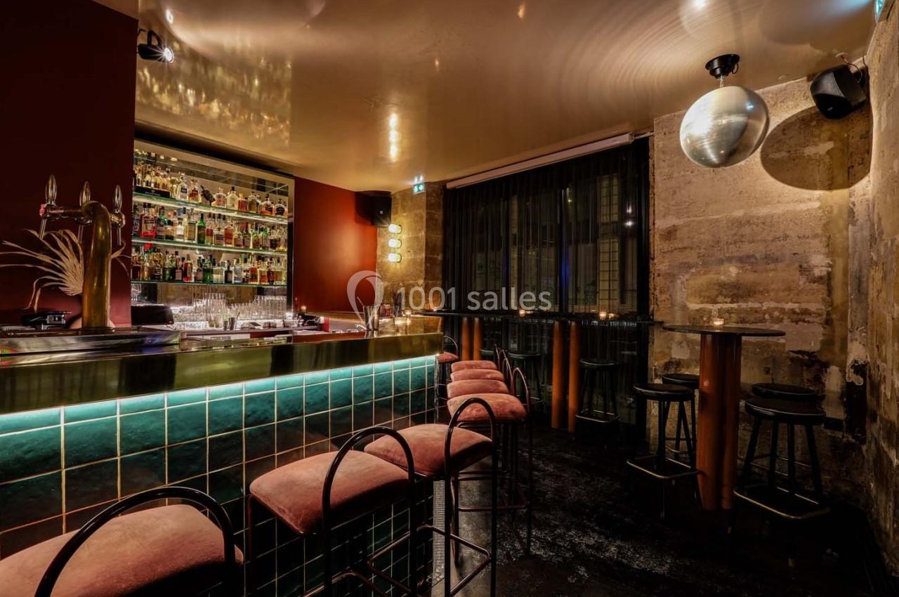 Intérieur d'un bar avec comptoir éclairé, tabourets rembourrés, tables hautes et mur en pierre apparente.