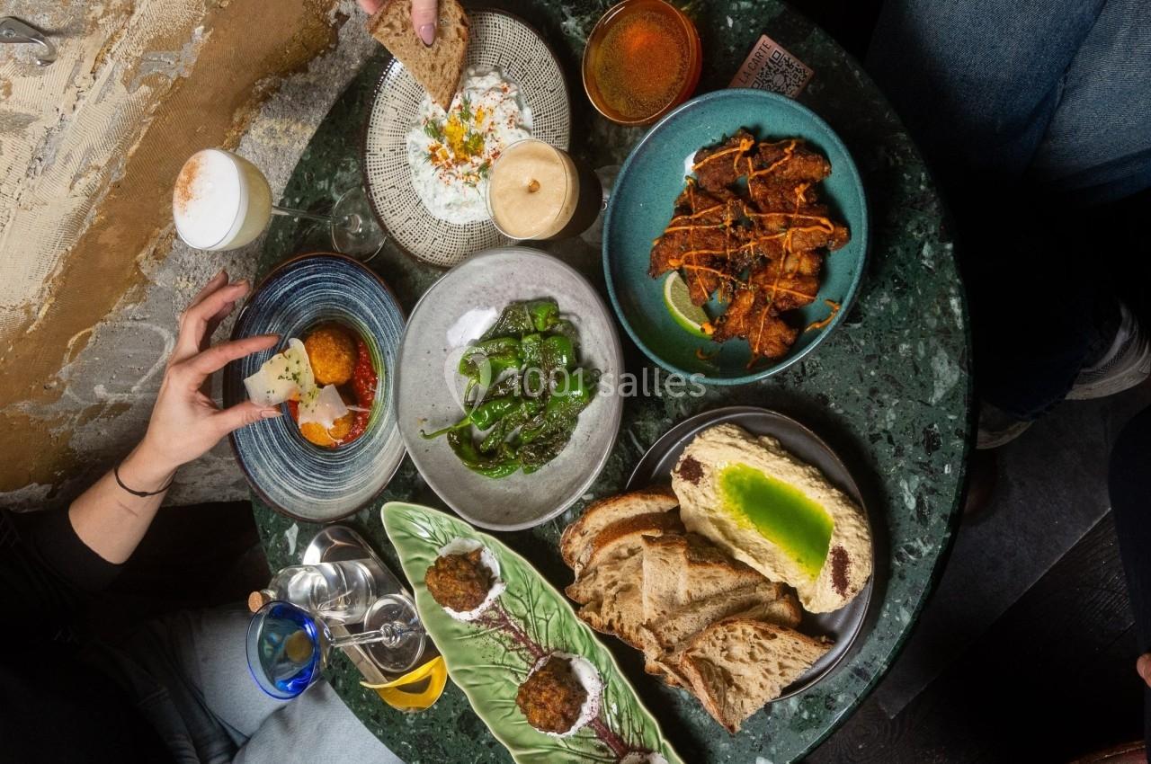 Table garnie de plats variés : pain, légumes, boulettes, sauces et boissons, avec des mains en train de servir.