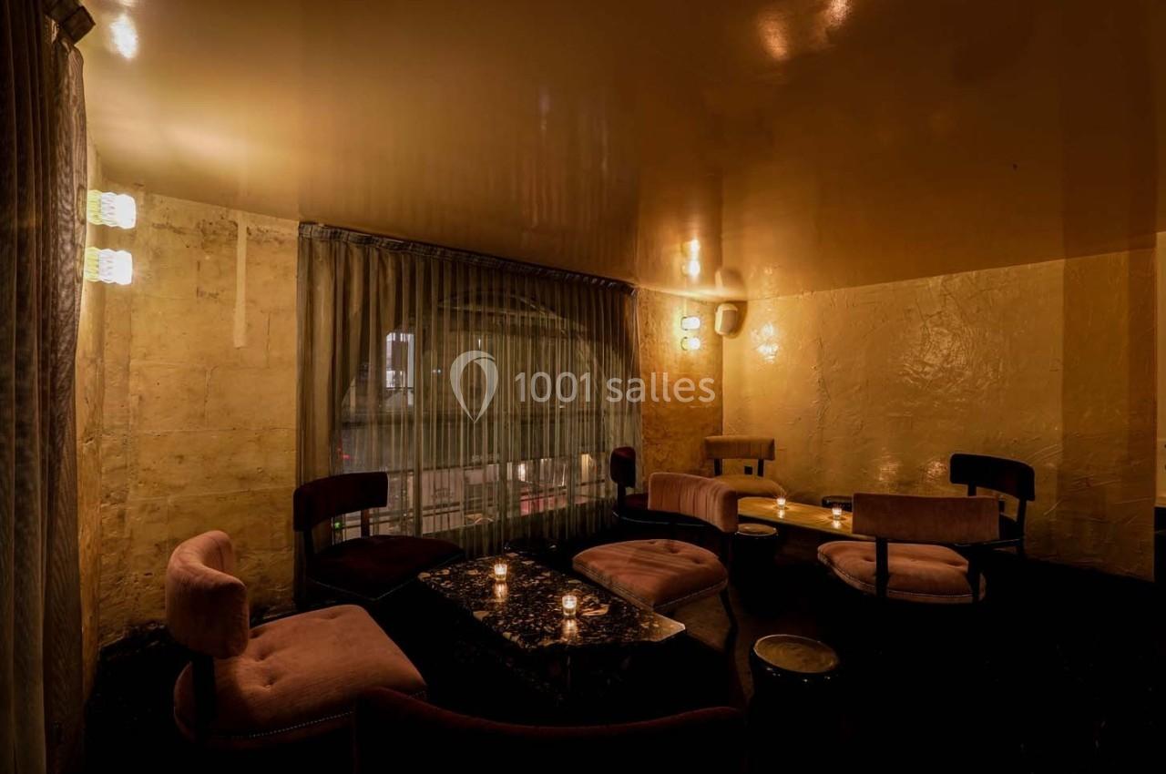 Salle intime avec éclairage tamisé, fauteuils en velours rose, table basse et rideaux donnant sur une fenêtre.