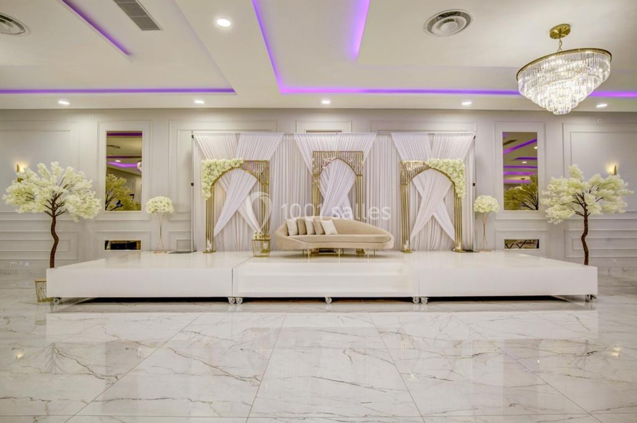 Estrade décorée avec un canapé beige, arches dorées, rideaux blancs, arbres artificiels et éclairage violet.