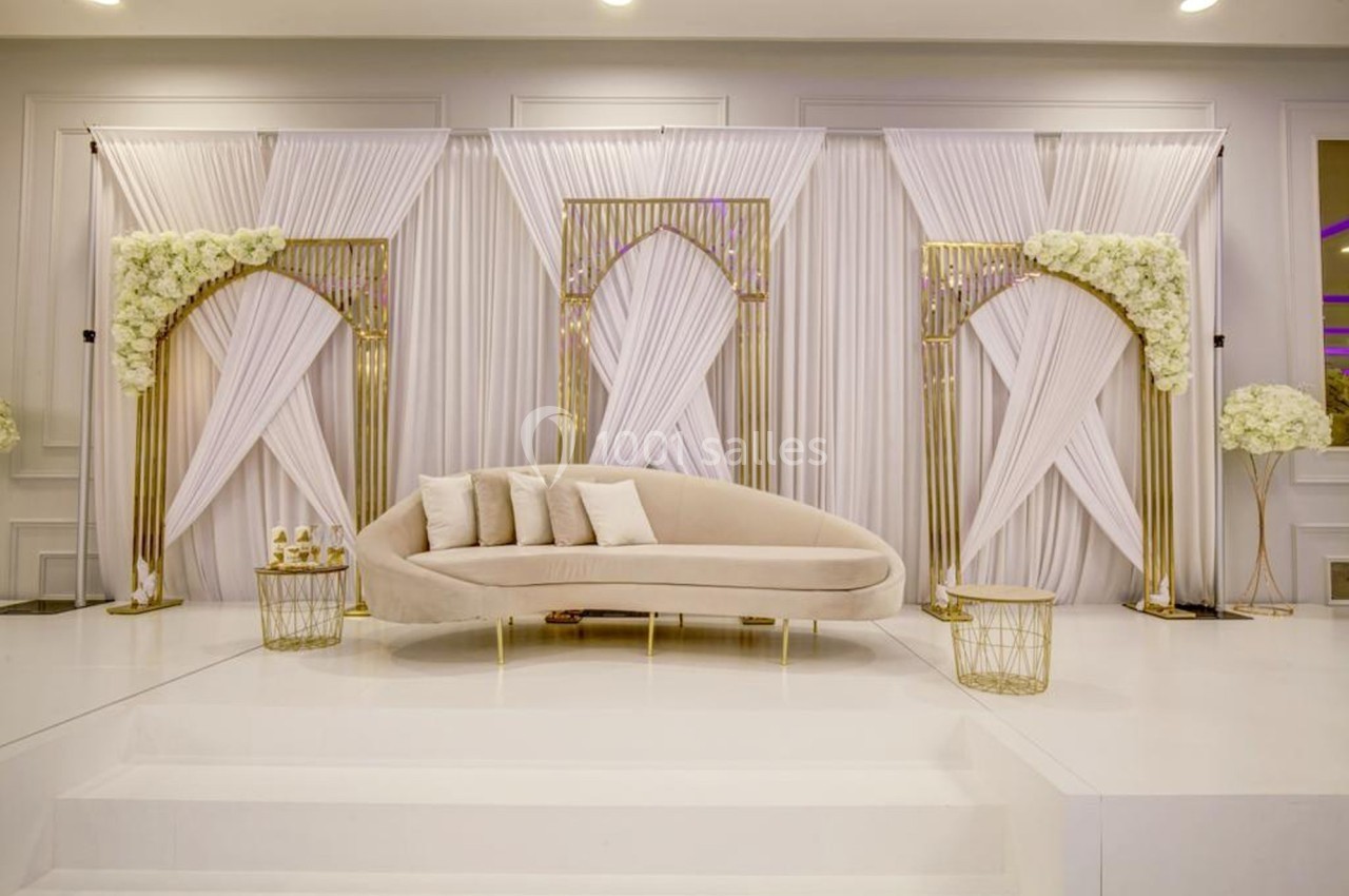 Canapé beige devant des rideaux blancs plissés et des arches dorées décorées de fleurs blanches.