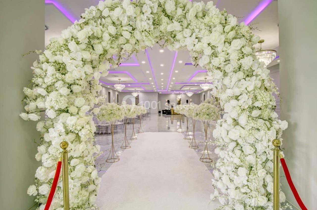 Arche de fleurs blanches décorant une allée centrale dans une salle lumineuse avec éclairage violet au plafond.