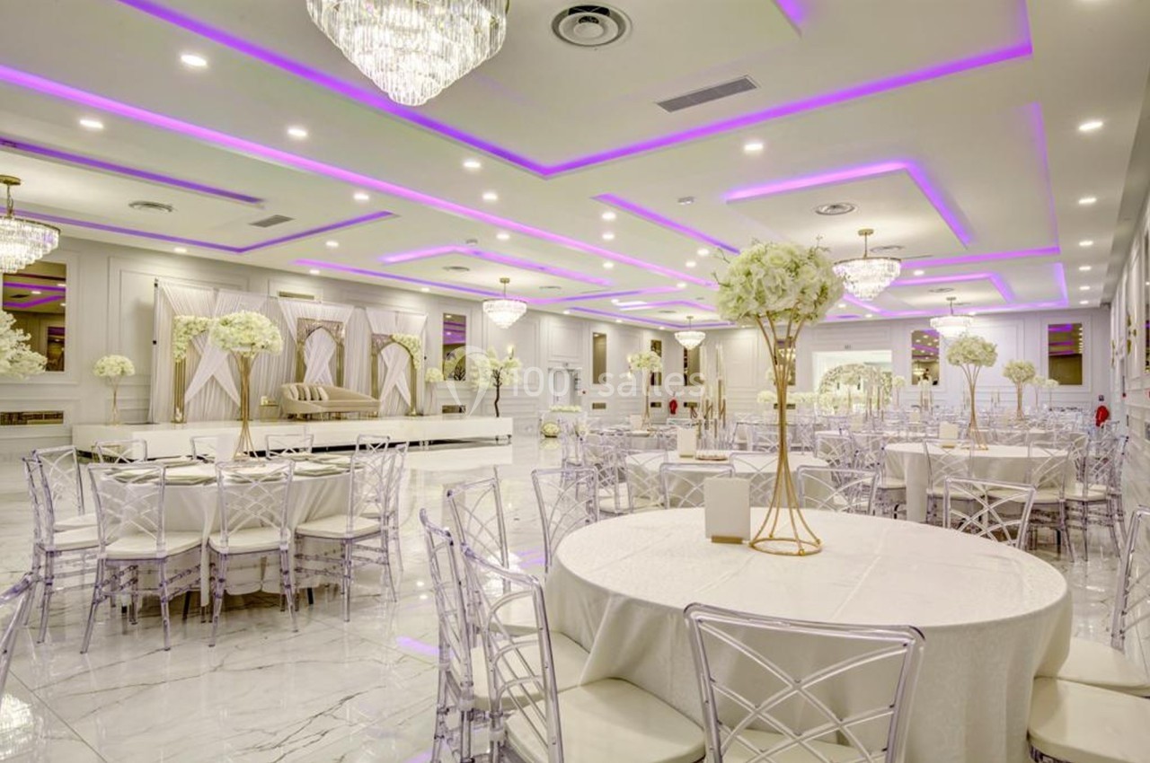 Salle de réception élégante avec tables rondes, chaises transparentes, décorations florales et éclairage violet au plafond.