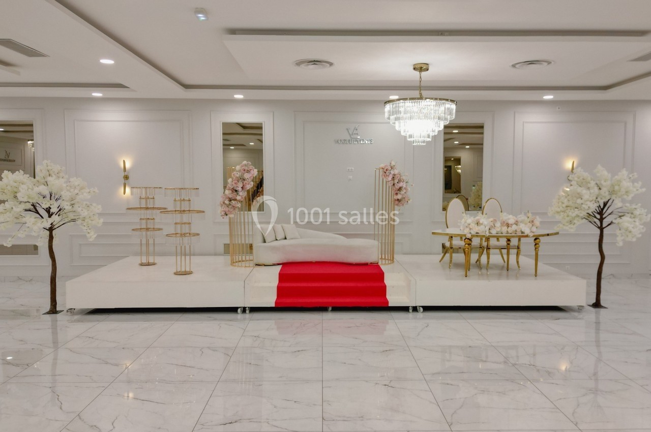 Salle de réception élégante avec une scène décorée de fleurs blanches et roses, un canapé et un tapis rouge.