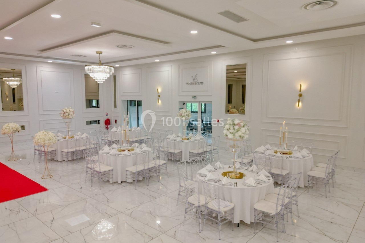 Salle de réception élégante avec tables rondes décorées de fleurs blanches, chaises transparentes et lustres suspendus.