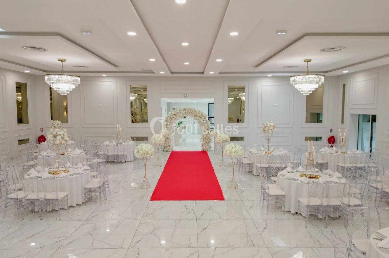 Salle de réception élégante avec tables décorées, arche florale blanche et tapis rouge au centre.