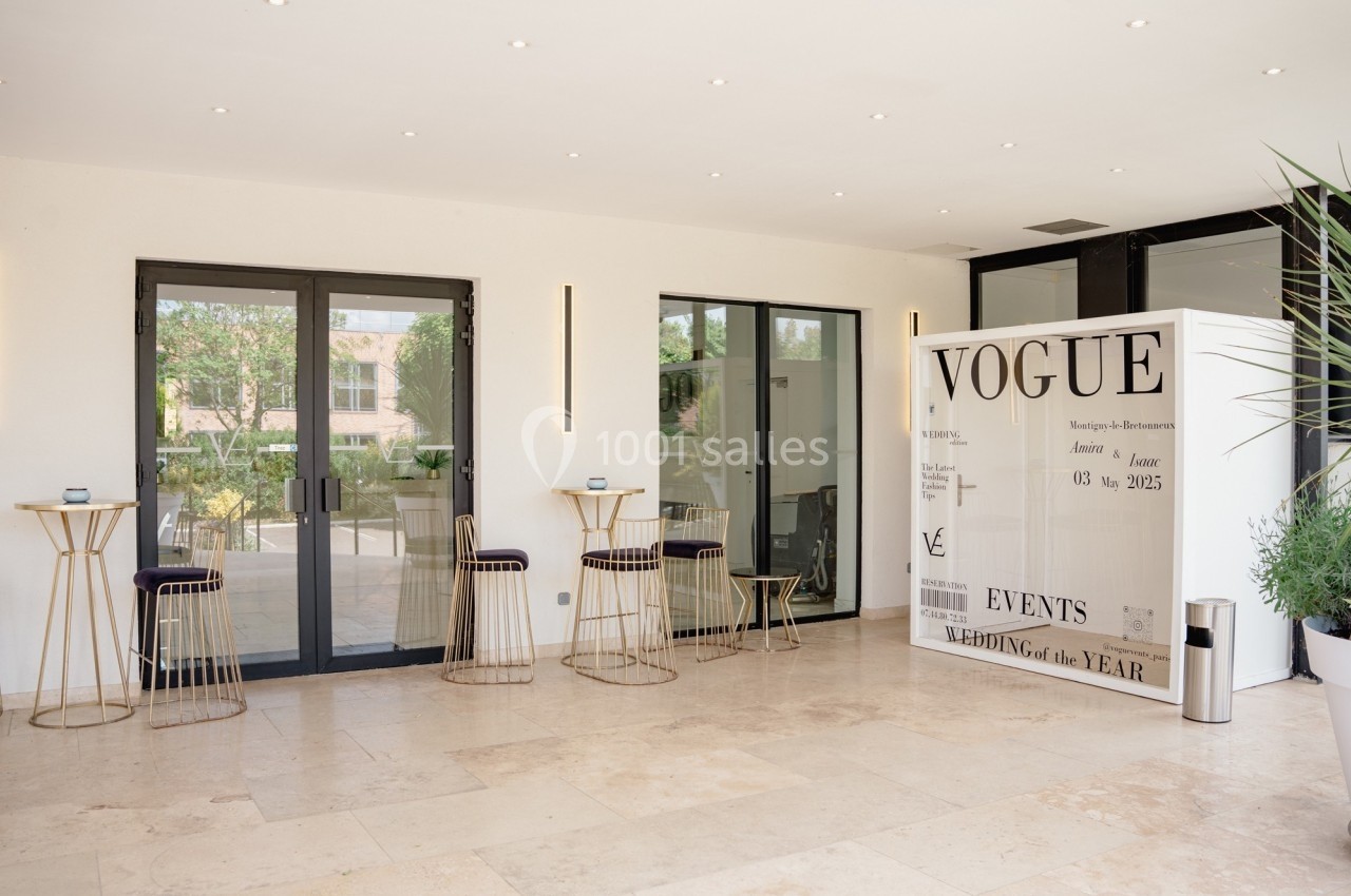 Salle lumineuse avec sol en pierre, tables hautes, chaises noires et une cabine photo blanche marquée ’Vogue’.