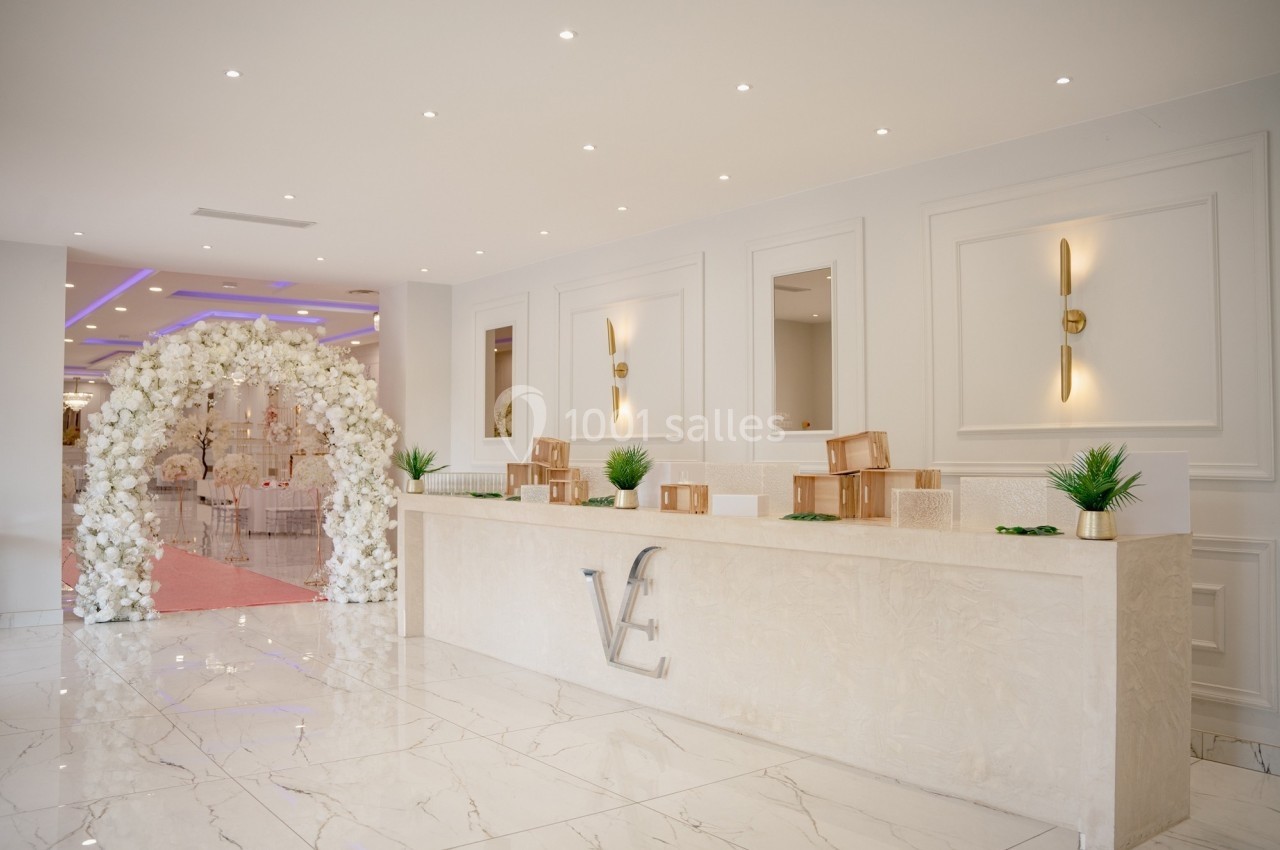 Comptoir d'accueil élégant dans une salle de réception, décoré de plantes et donnant sur une arche florale blanche.