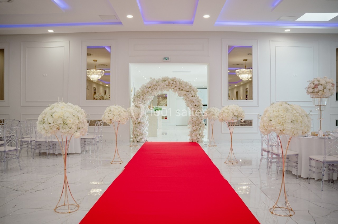 Salle de réception élégante avec arche florale blanche, tapis rouge et tables décorées de bouquets de fleurs blanches.
