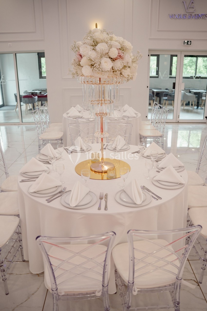 Table décorée avec nappes blanches, serviettes pliées, vaisselle élégante et centre de table floral sur pied doré.