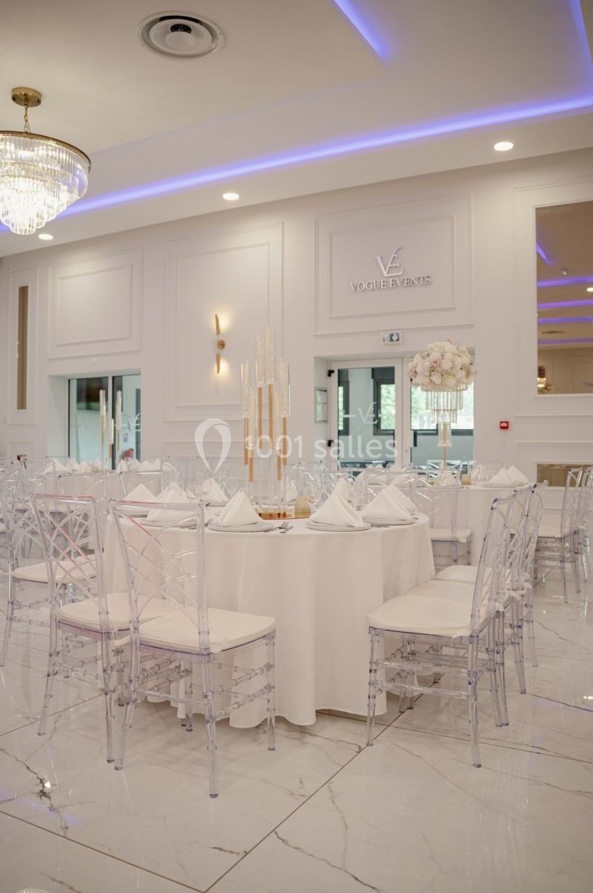 Salle de réception élégante avec tables rondes dressées, chaises transparentes et décorations raffinées.