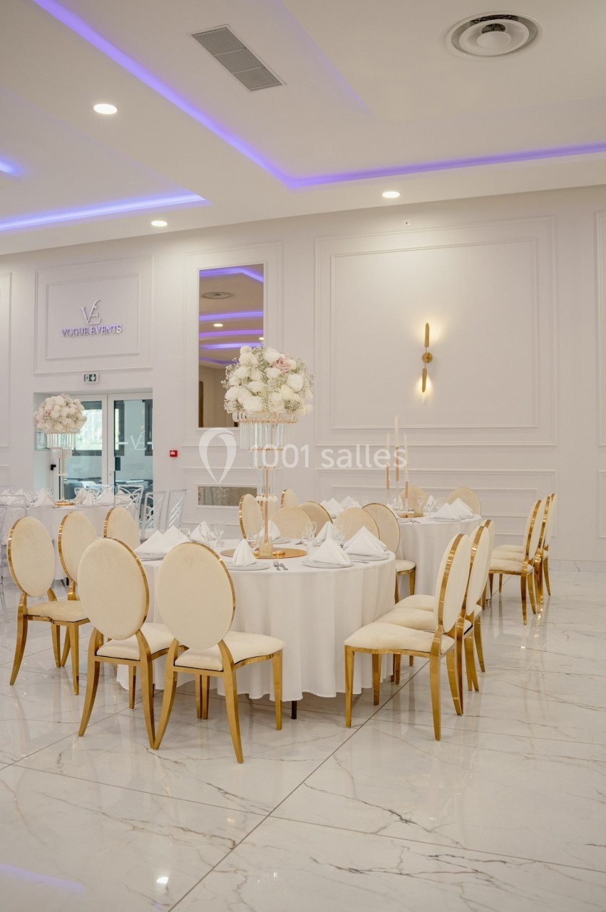 Salle de réception élégante avec tables rondes dressées, chaises dorées et décoration florale blanche.