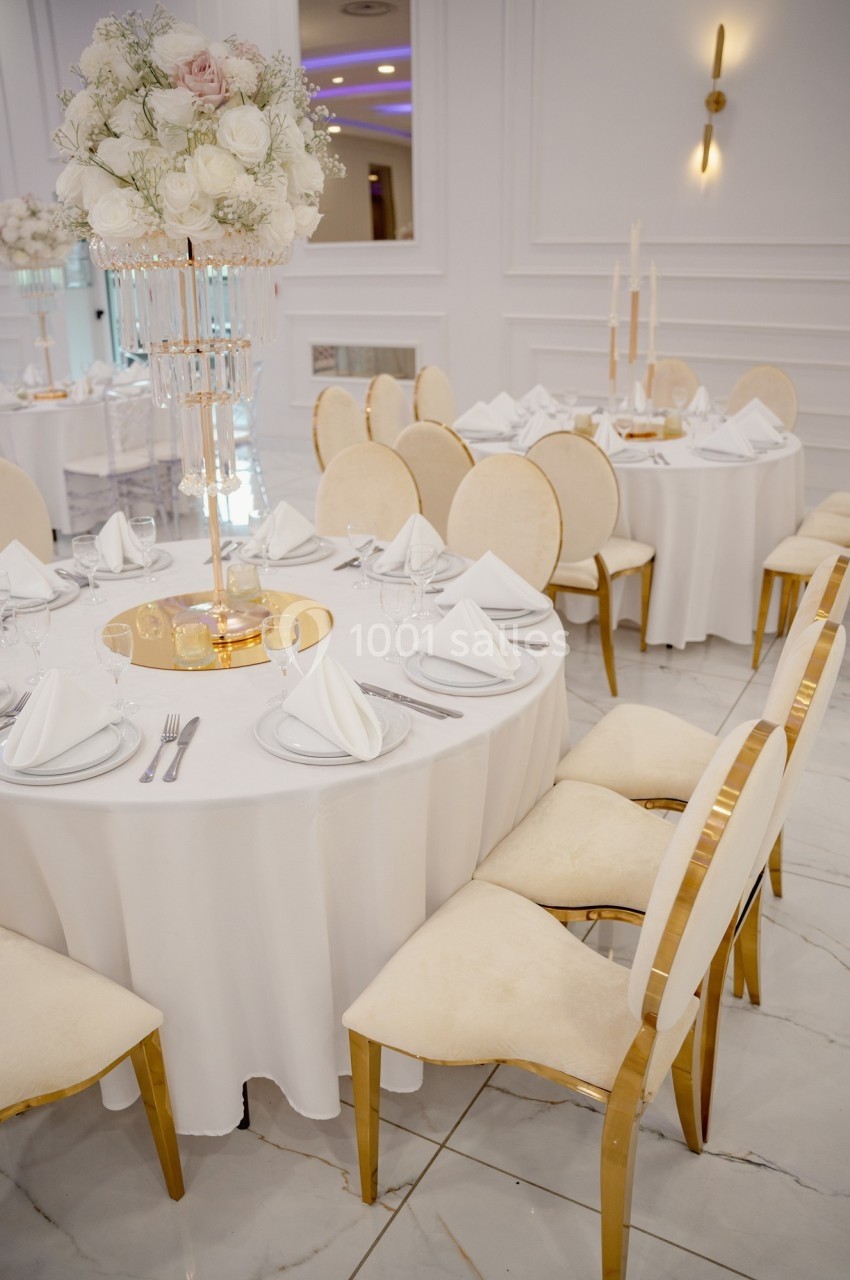 Salle de réception élégante avec tables rondes dressées, nappes blanches, chaises dorées et centre de table floral.