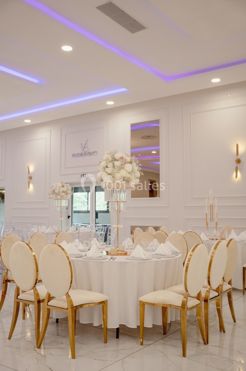 Salle de réception élégante avec tables rondes dressées, chaises dorées et centre de table floral blanc.