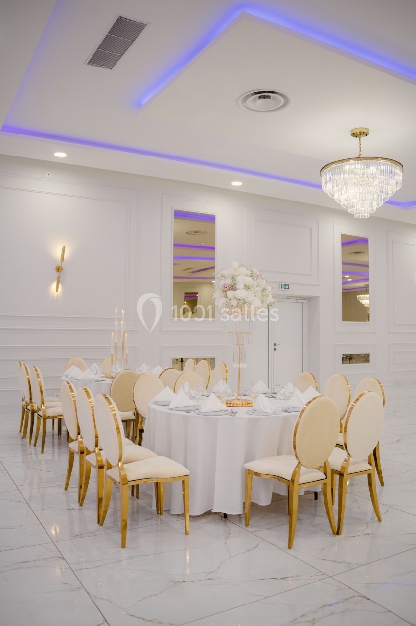Salle de réception élégante avec table ronde dressée, chaises dorées, décoration florale et éclairage raffiné.