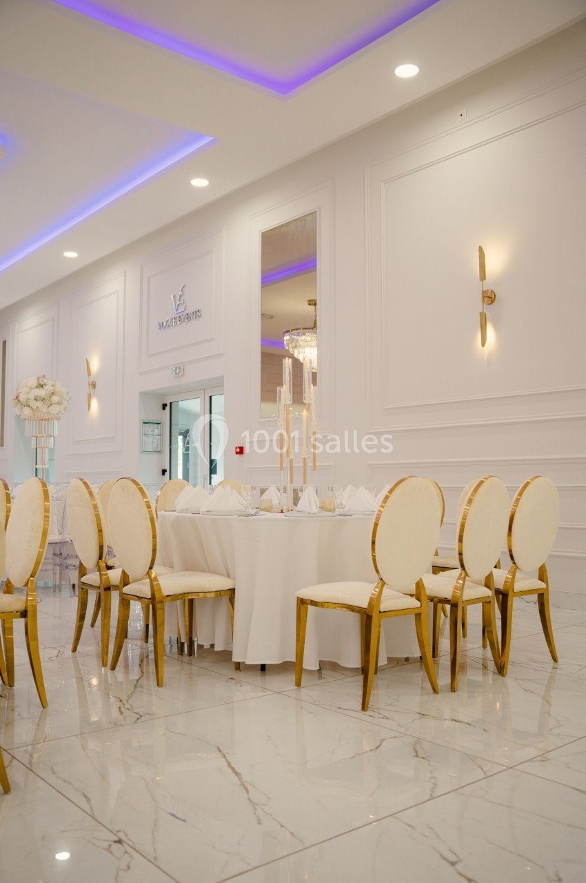 Salle de réception élégante avec table ronde dressée, chaises dorées et éclairage moderne sur un sol en marbre.