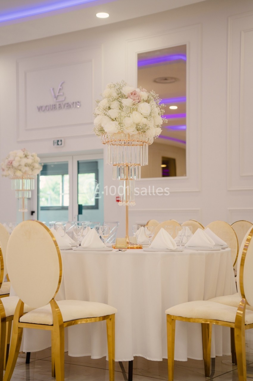 Table décorée pour un événement, avec nappes blanches, chaises dorées et centre de table floral élégant.