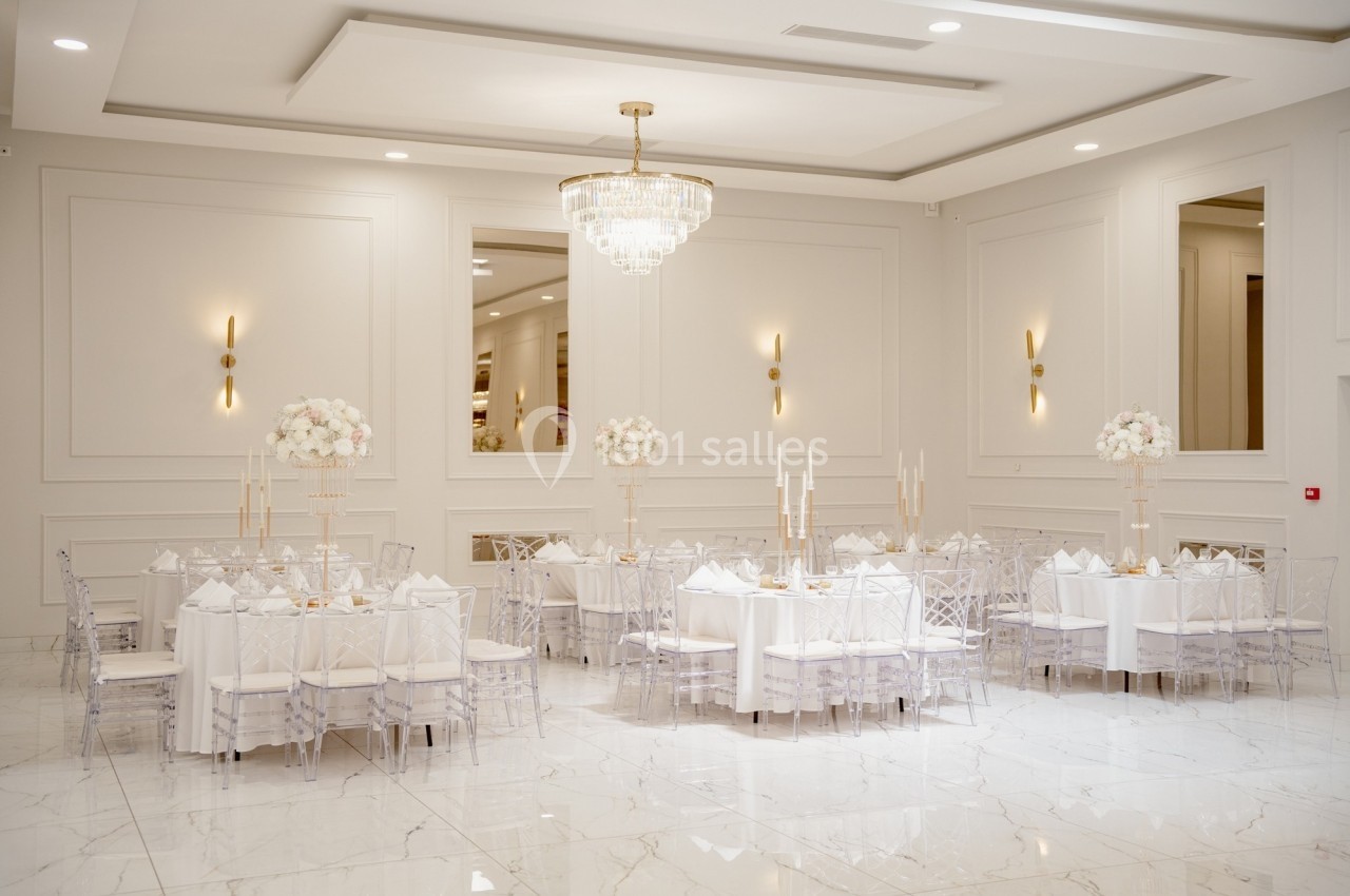 Salle de réception élégante avec tables décorées de fleurs blanches, chaises transparentes et lustre central.