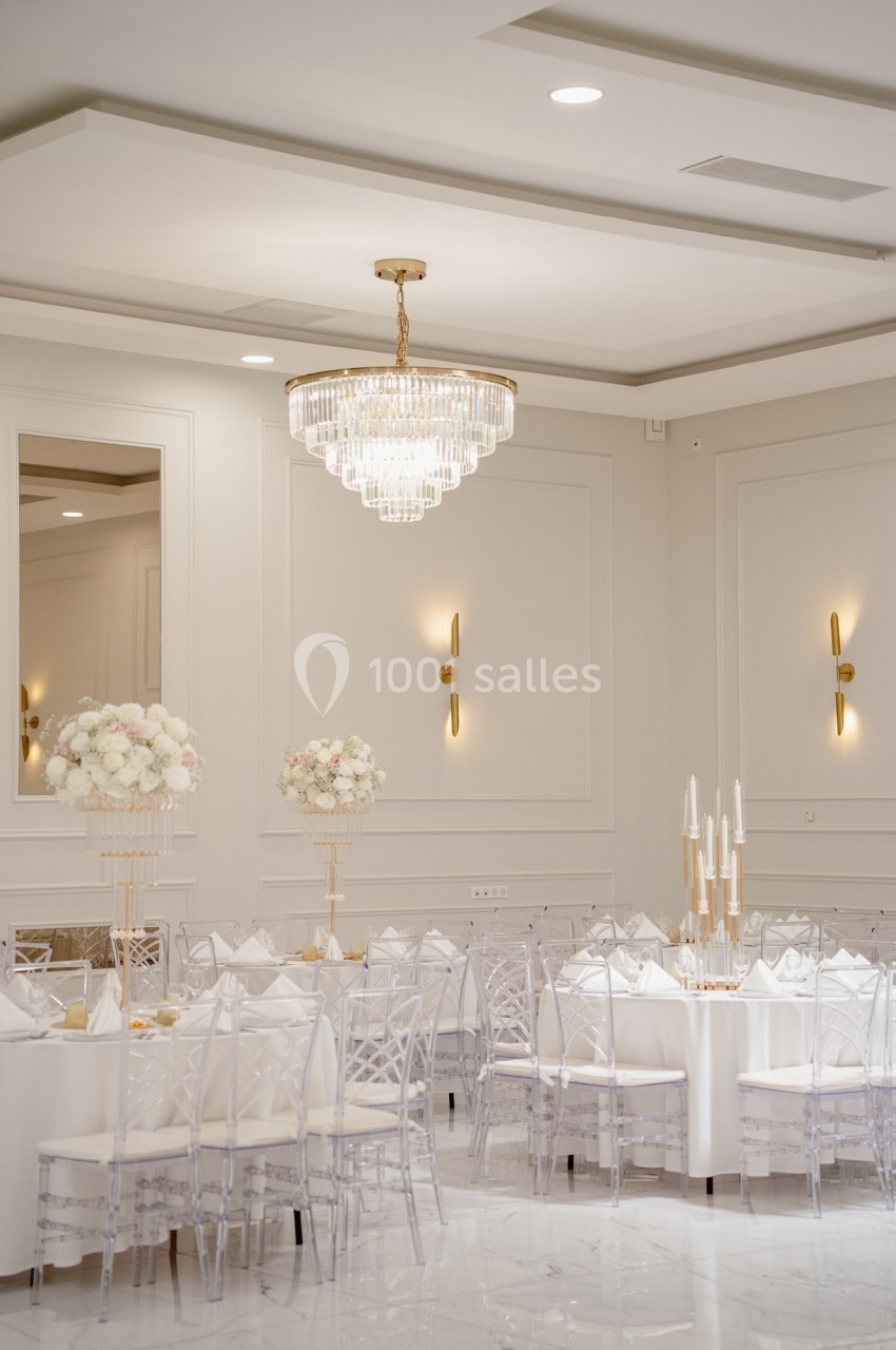 Salle de réception élégante avec tables dressées, chaises transparentes, décorations florales blanches et lustre en cristal.