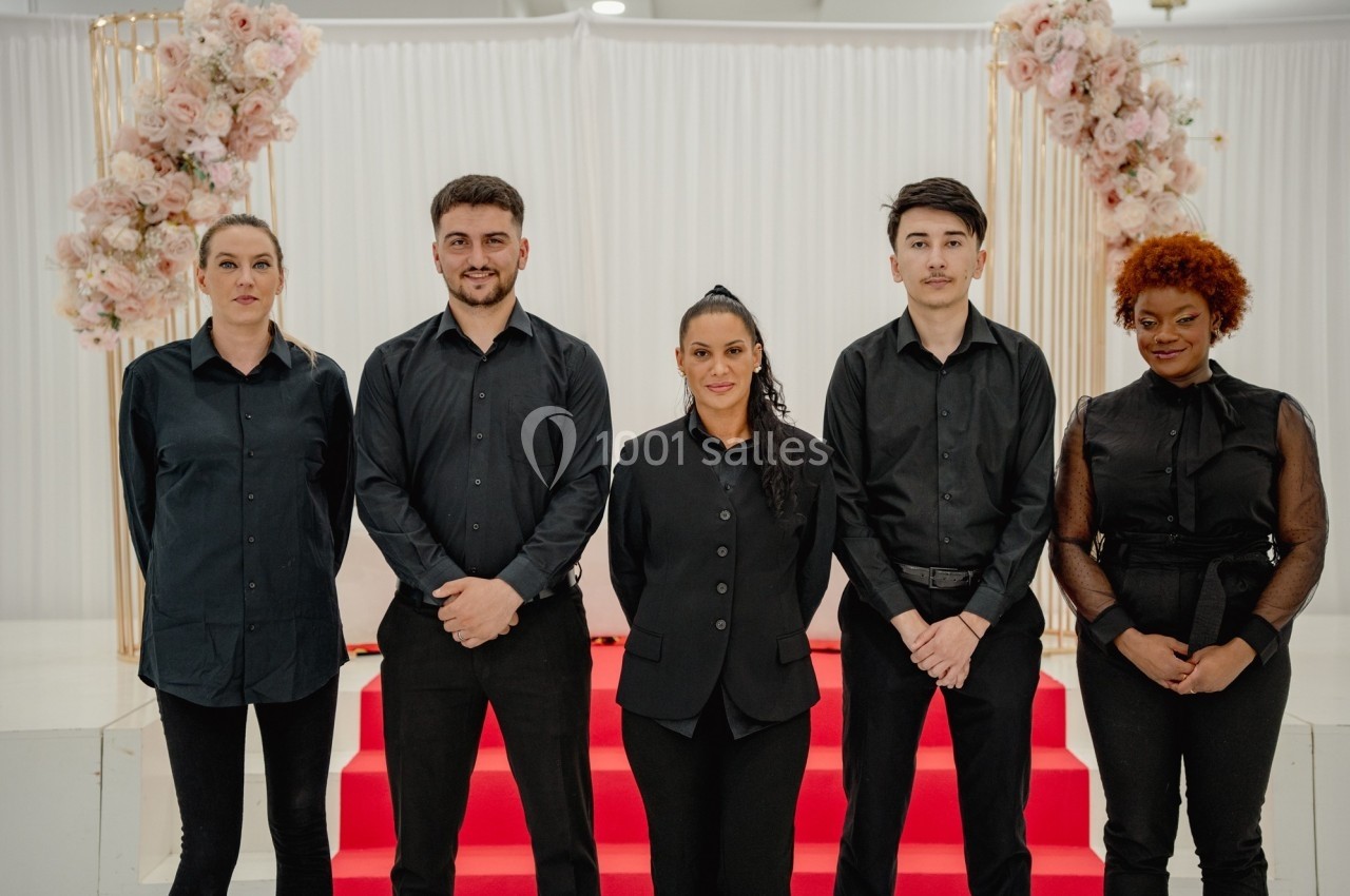 Cinq personnes en tenue noire posent debout devant un décor avec rideaux blancs, fleurs roses et marches rouges.