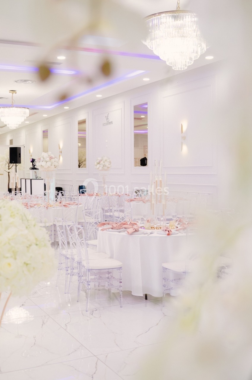 Salle de réception élégante avec tables rondes dressées, chaises transparentes et décorations florales blanches.