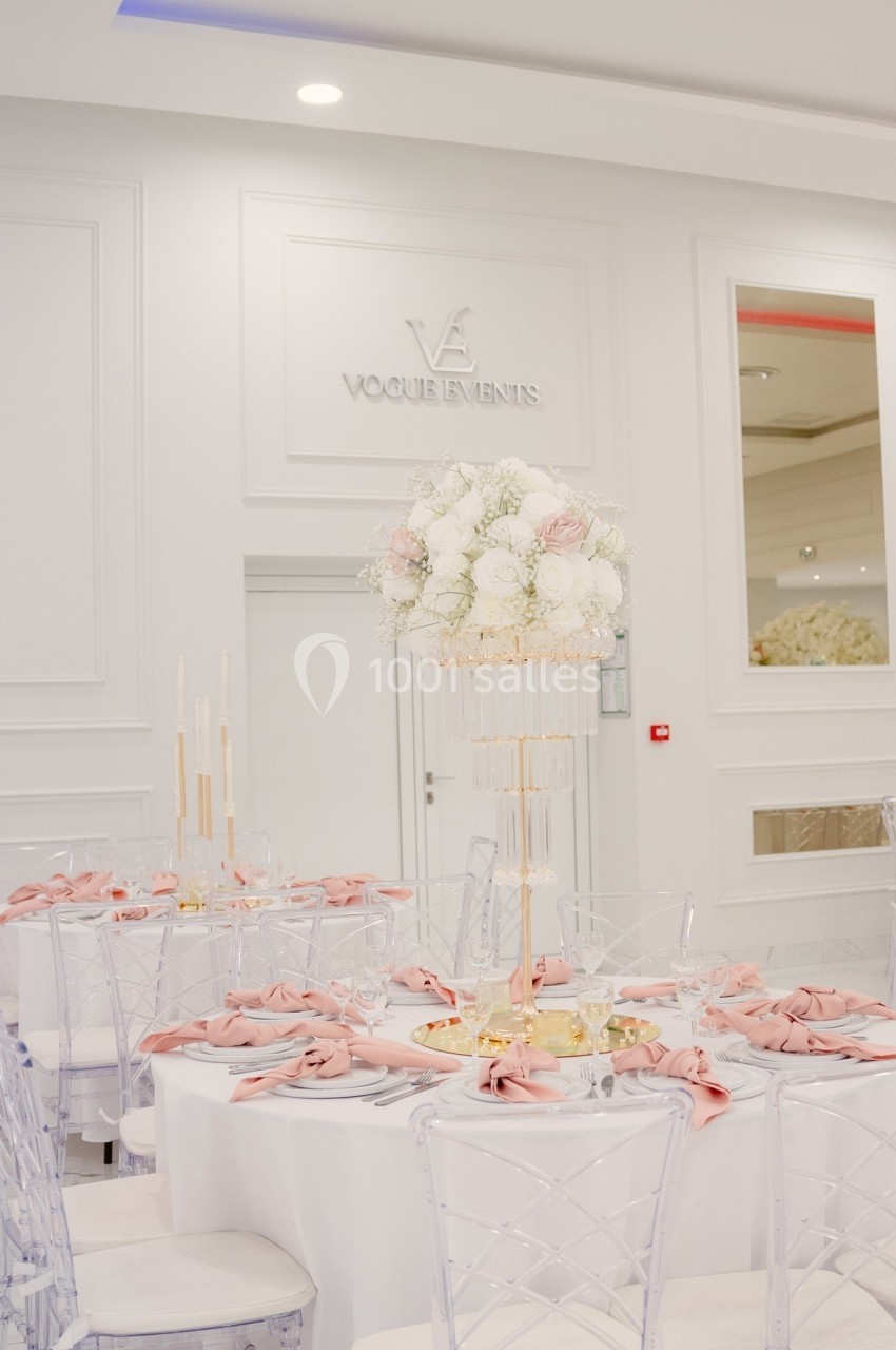 Salle de réception élégante avec table décorée de fleurs blanches et roses, serviettes roses et chaises transparentes.