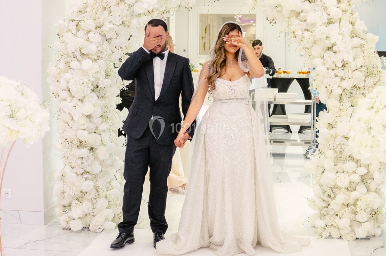 Un couple en tenue de mariage se tenant la main, les yeux couverts, devant une arche décorée de fleurs blanches.