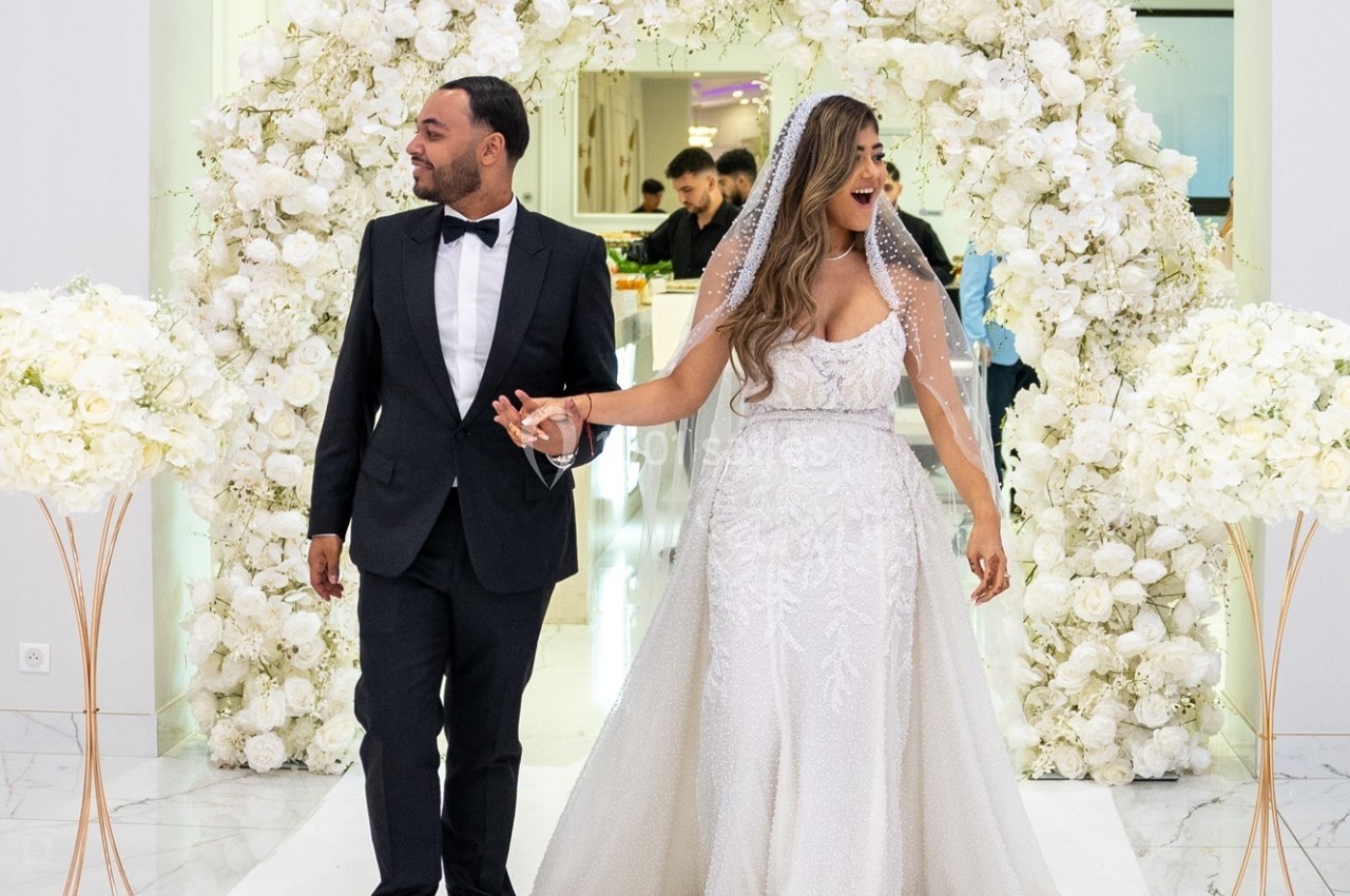 Un couple en tenue de mariage marche devant une arche florale blanche dans un décor élégant.