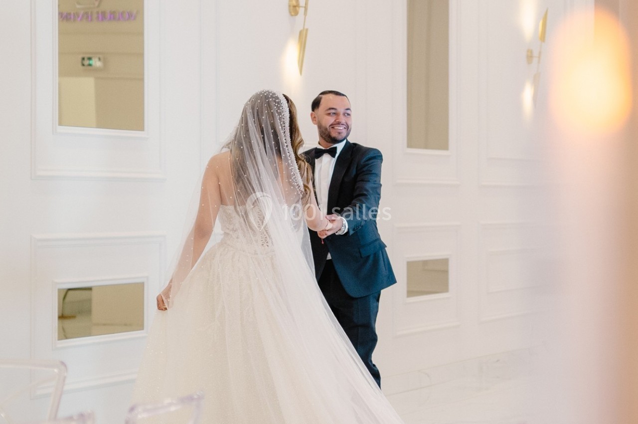 Un couple en tenue de mariage danse dans une salle lumineuse aux murs blancs et décorations élégantes.