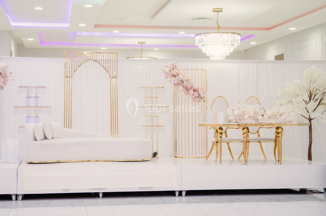 Salle décorée avec élégance, comprenant un canapé blanc, une table dorée ornée de fleurs et un éclairage raffiné.