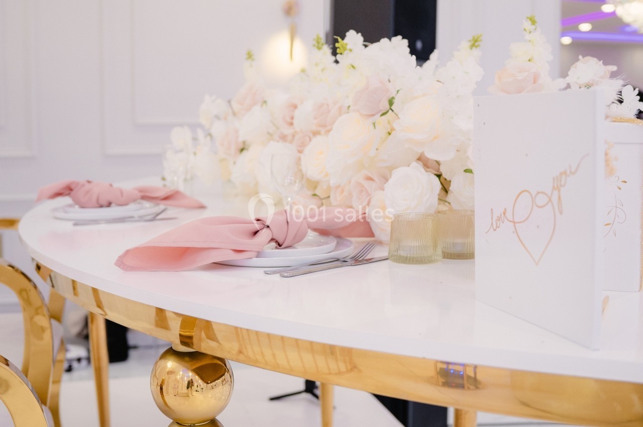 Table élégante décorée de fleurs blanches et roses, serviettes roses pliées et menus posés sur une surface blanche brillante.