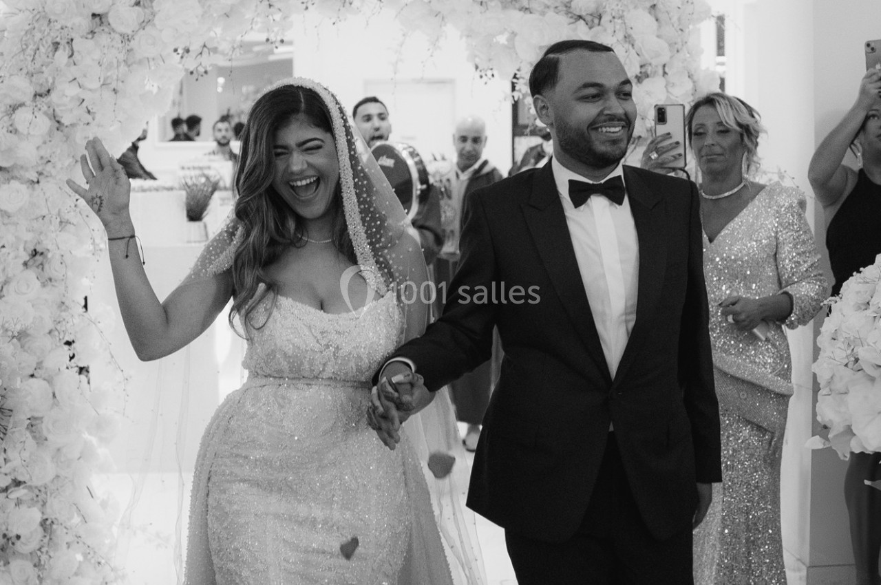 Un couple souriant marche sous une arche florale lors d'une cérémonie de mariage, entouré d'invités.