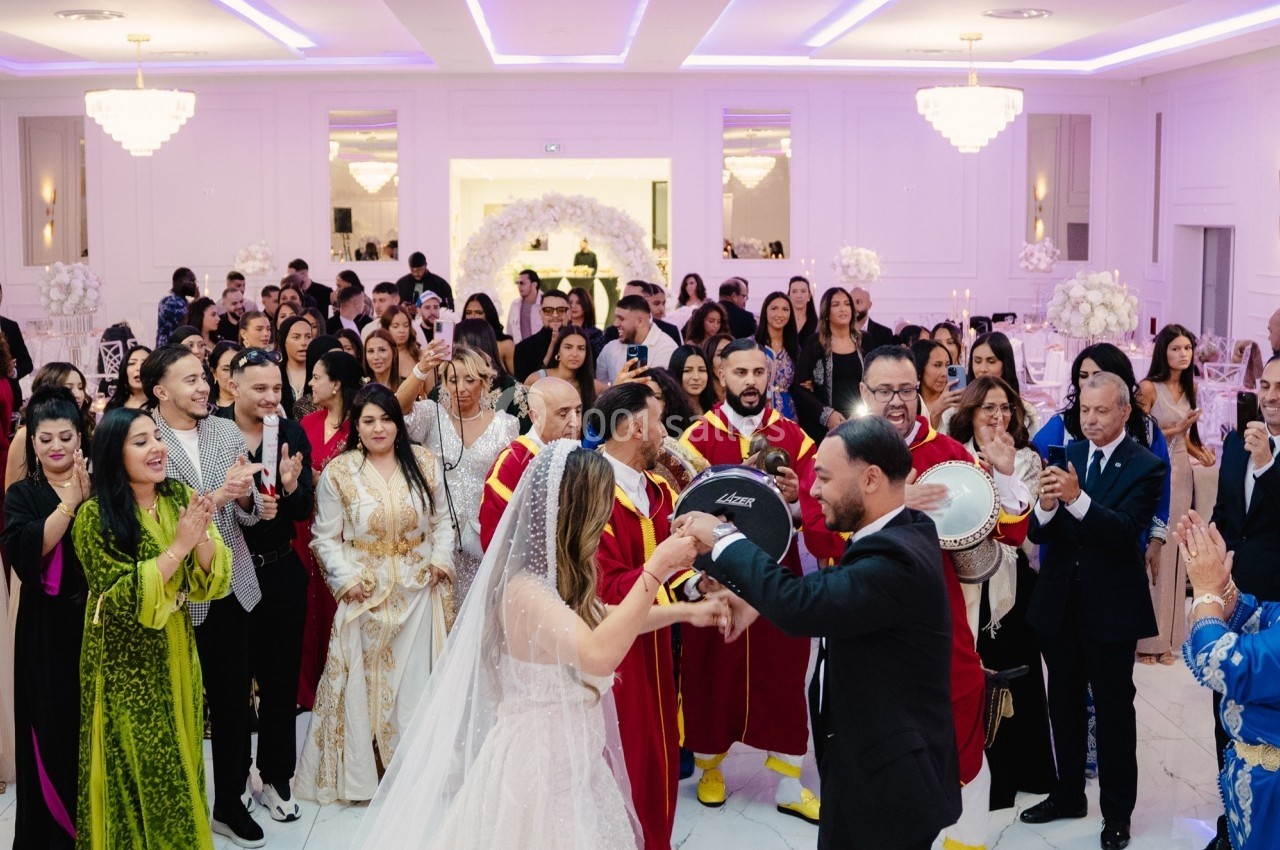 Un couple en tenue de mariage danse entouré d'invités applaudissant dans une salle décorée de lumières et de fleurs blanches.