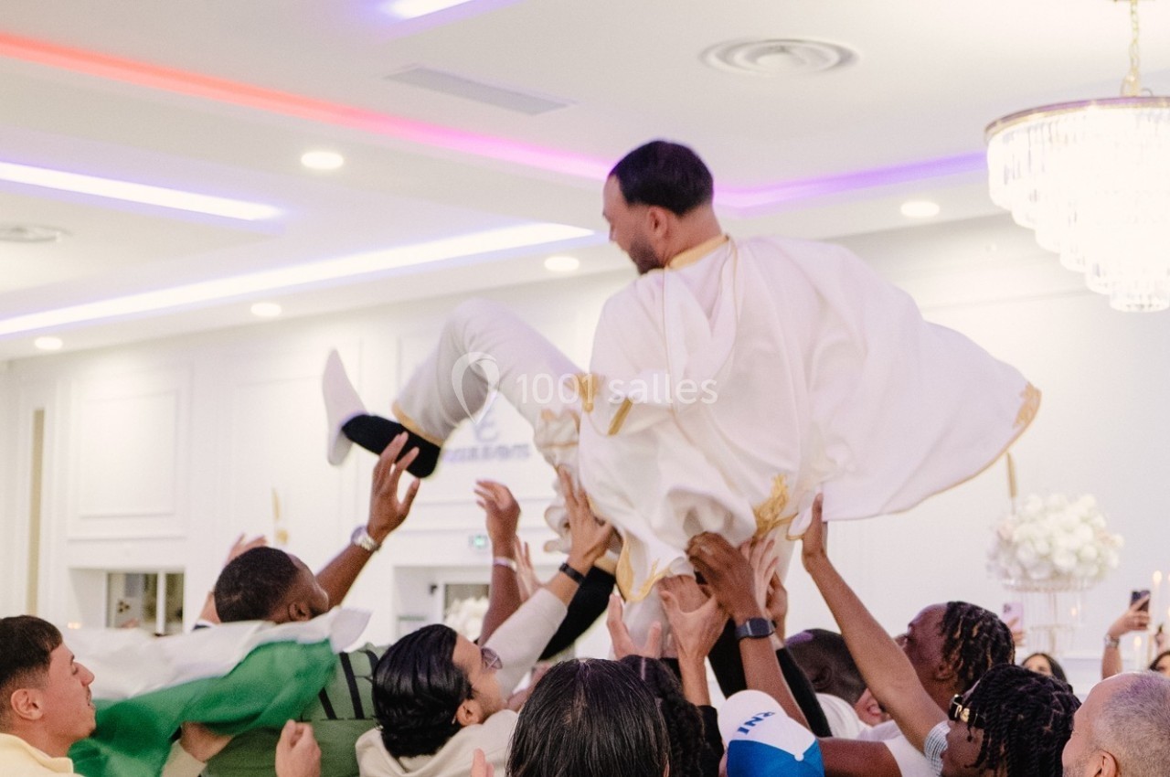 Un homme en tenue blanche traditionnelle est soulevé en l'air par un groupe de personnes dans une salle festive éclairée.