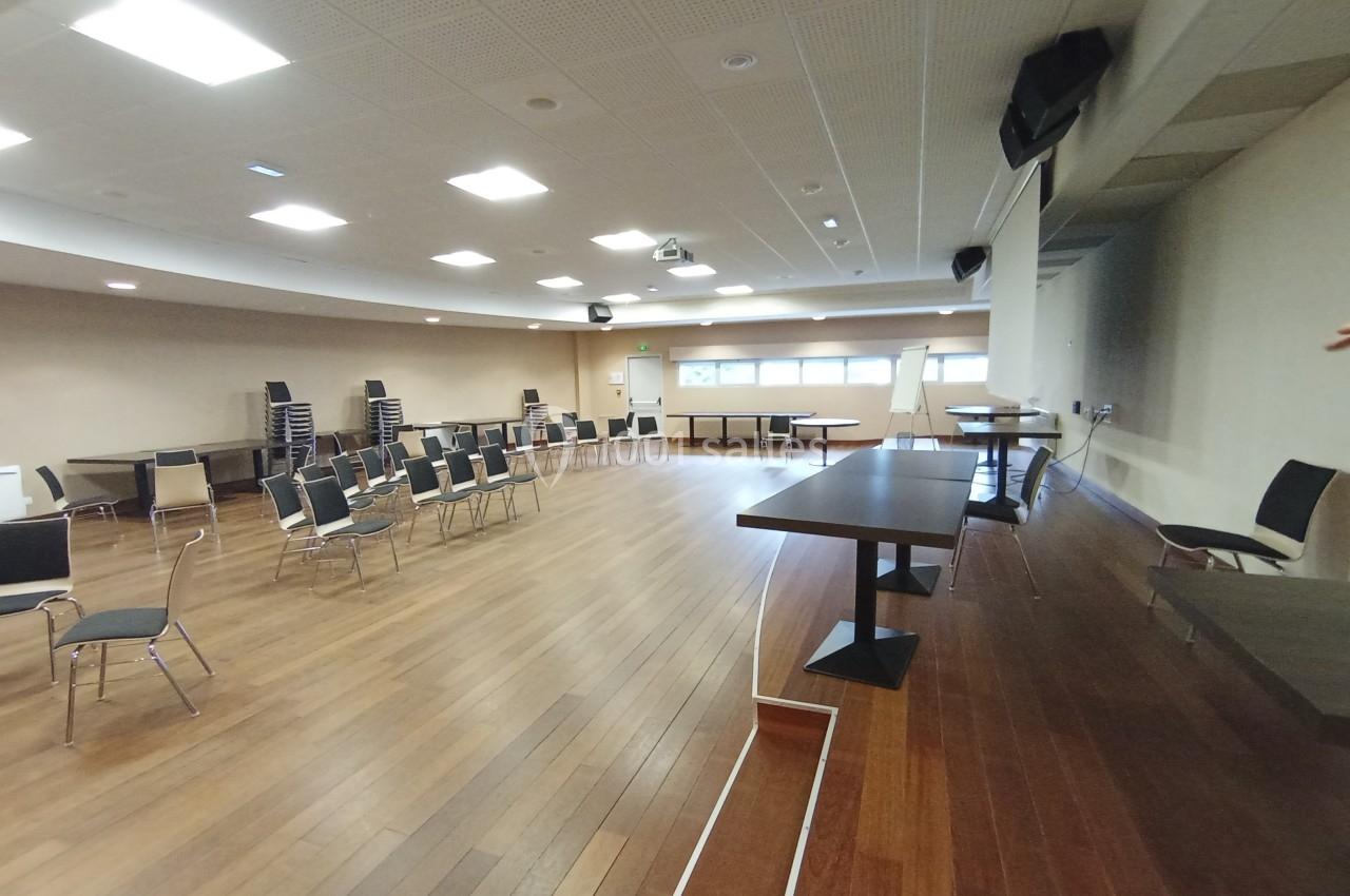 Salle vide avec parquet, chaises empilées et disposées, tables alignées, éclairage au plafond et estrade à droite.