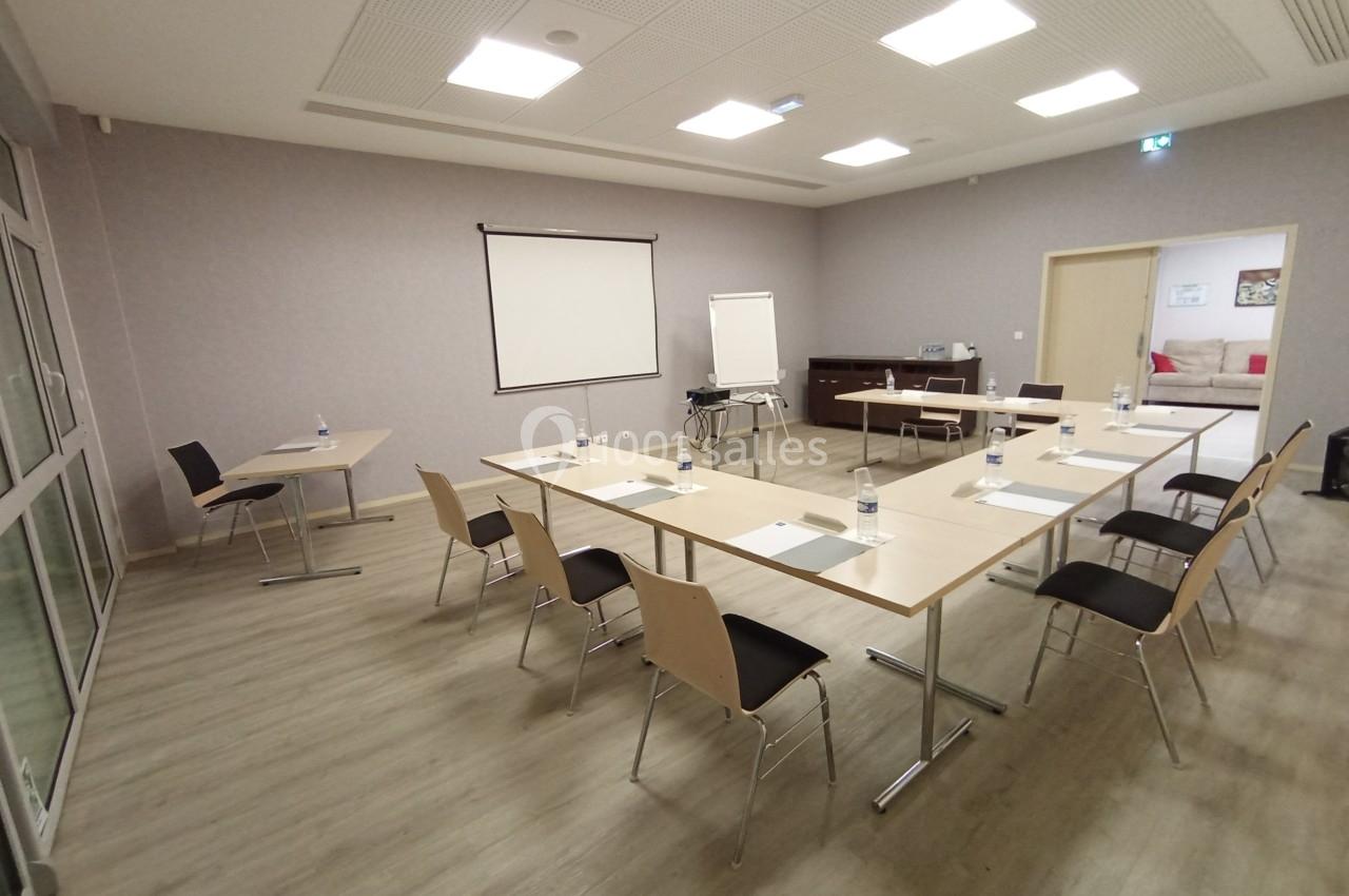 Salle de réunion lumineuse avec tables disposées en U, chaises, tableau blanc et écran de projection.