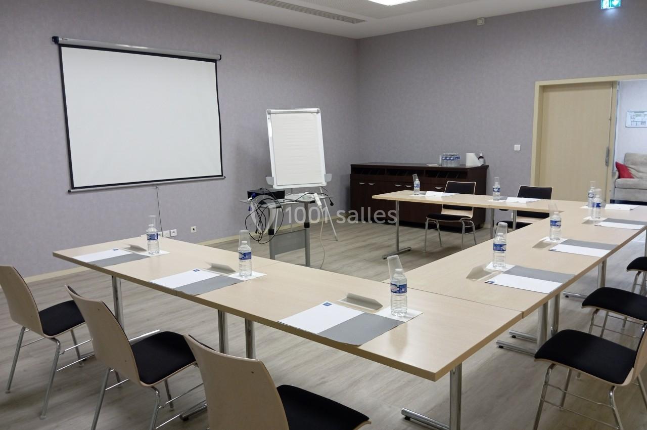 Salle de réunion équipée de tables disposées en U, chaises, paperboard, écran de projection et bouteilles d'eau.