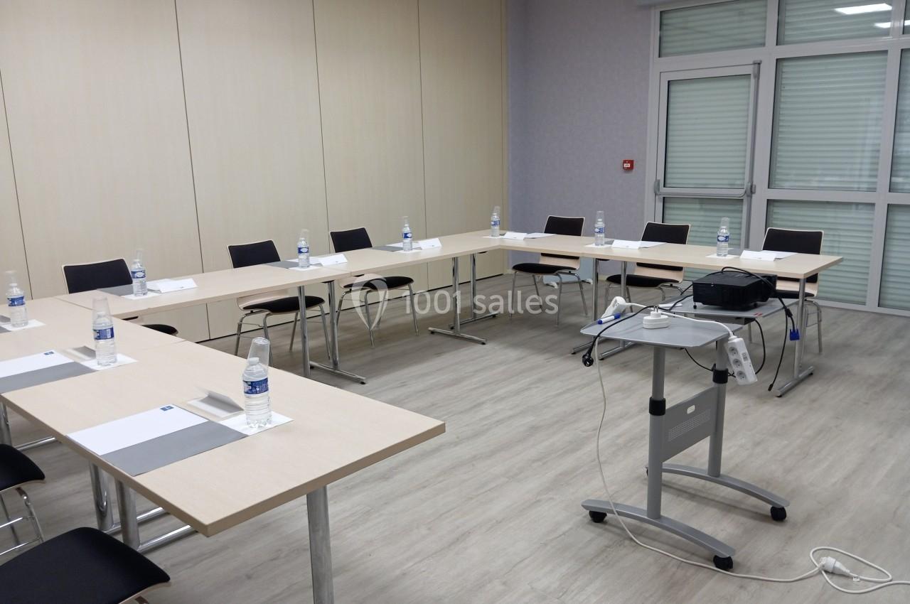 Salle de réunion avec tables disposées en U, chaises, bouteilles d'eau, documents et un projecteur sur support mobile.